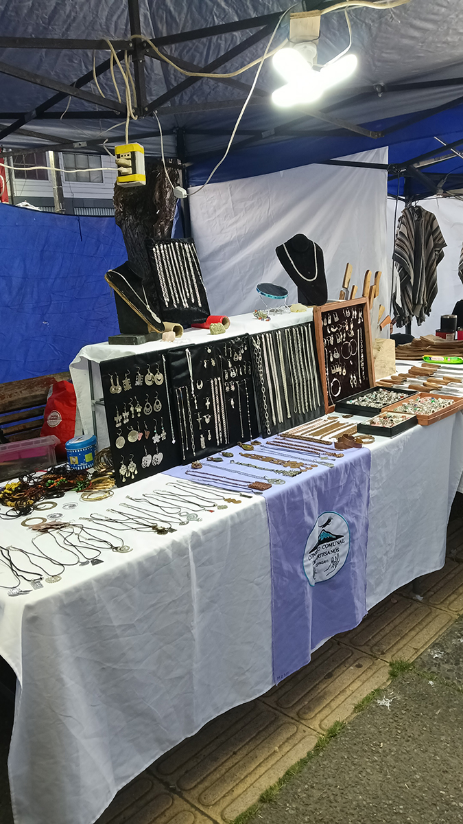 Puesto de feria artesanal con mesa cubierta por mantel blanco donde se exhiben collares, pulseras y aros organizados en paneles negros y bandejas, bajo una carpa iluminada.