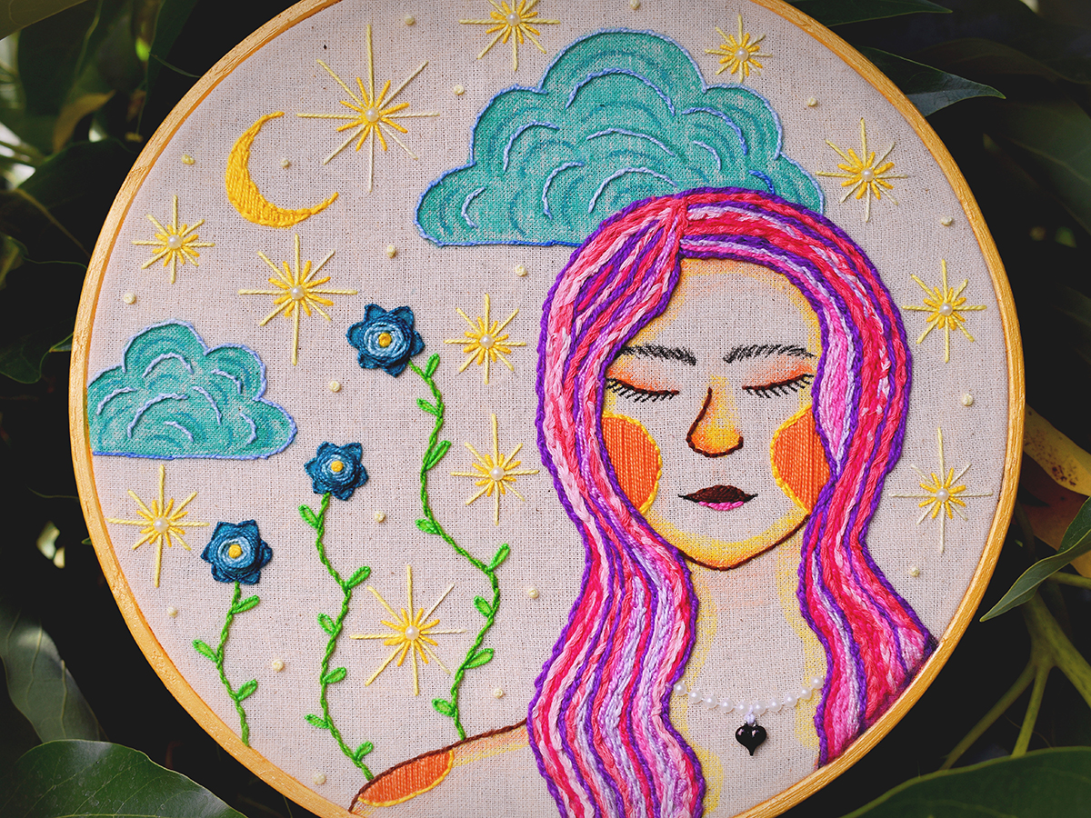 Bordado circular que representa el rostro de una mujer de ojos cerrados, con cabello rosado y morado, rodeada de estrellas, luna y flores sobre fondo claro.