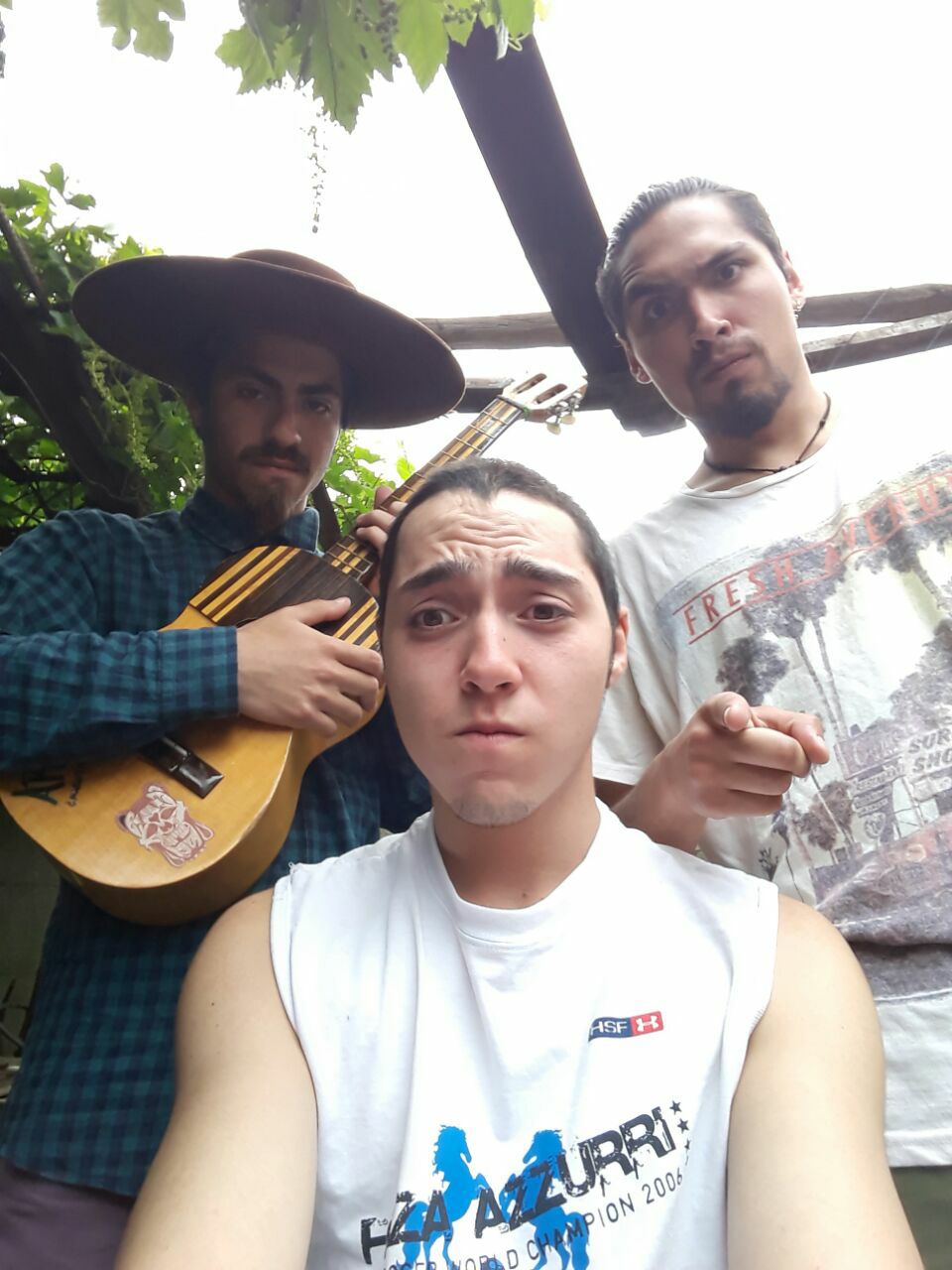 Tres jóvenes posan para una selfie al aire libre; uno sostiene una guitarra pequeña y viste sombrero amplio, mientras los otros miran a cámara con expresión seria bajo una parra.